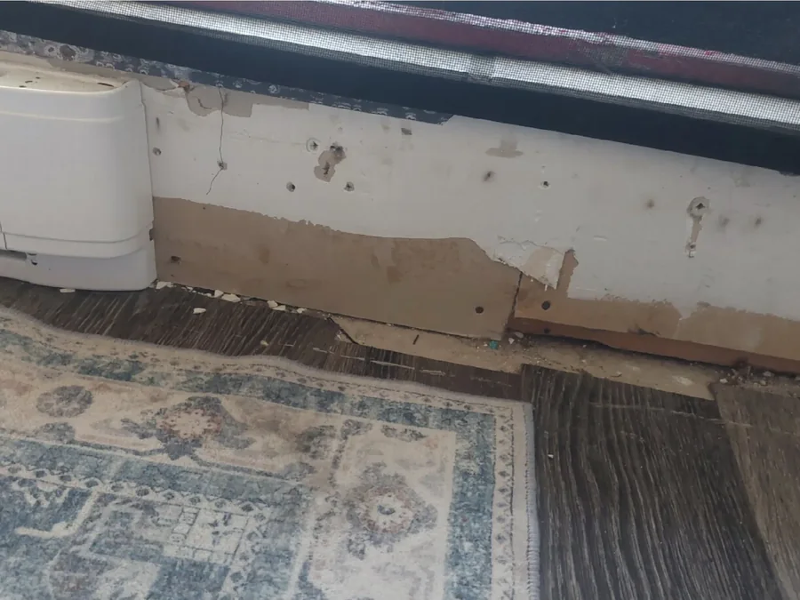 Wall torn open behind a baseboard heater, with damaged flooring